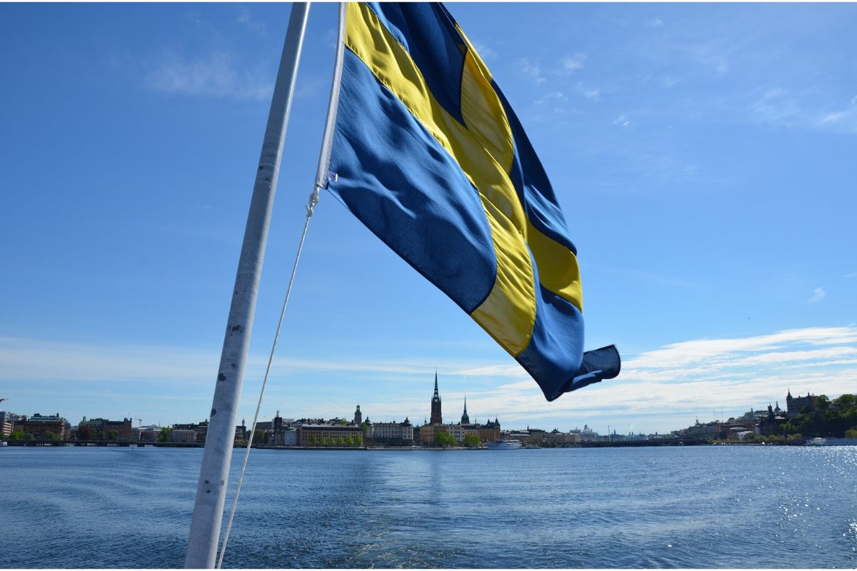 Powiewająca flaga szwedzka nad powierzchnią wody, a na horyzoncie budynki Sztokholmu
